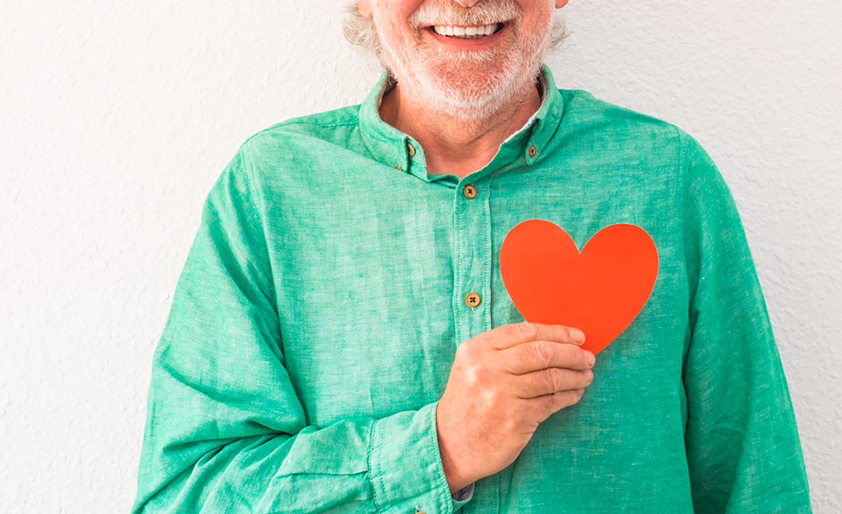 Colourful posed photo of a smiling elderly man against a bright turqouise background, holding a red paper heart to his chest. 