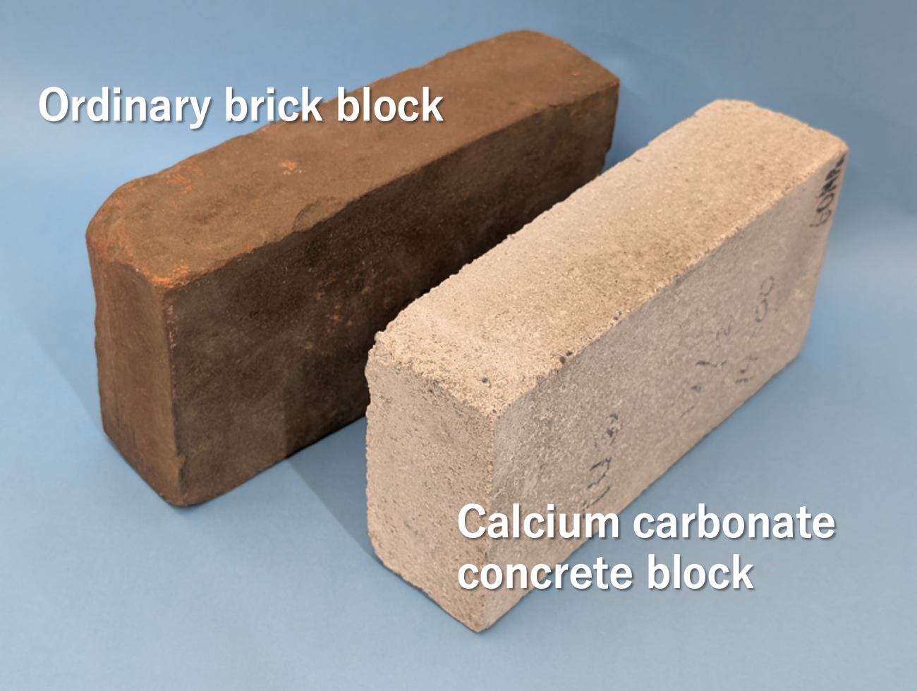  Photograph of two concrete blocks against a light blue background. The left is made of new concrete and is dark brown, the right is made of recycled concrete and is cream colored.