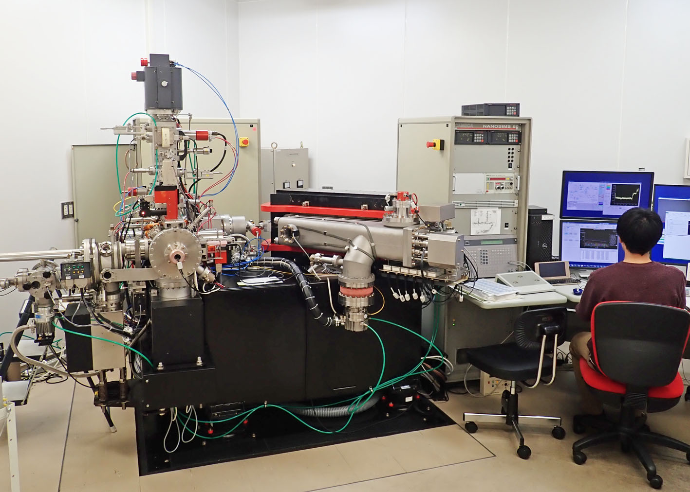 A person sitting at a large complicated machine of metal parts, tubes and wires