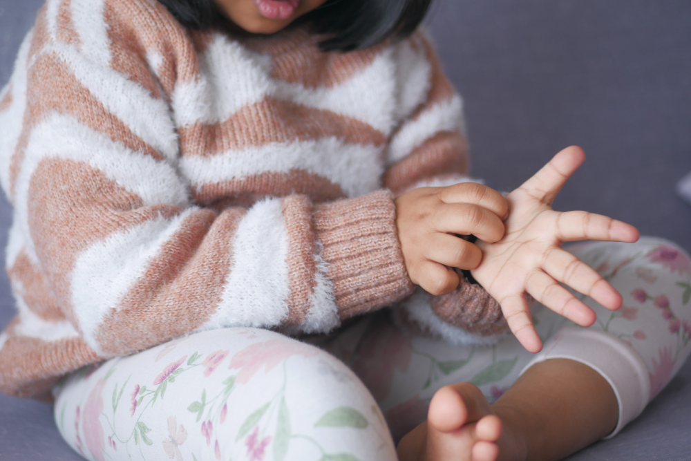 Photo of young girl with black hair in comfy clothes, sitting on the ground and scratching the palm of her hand.